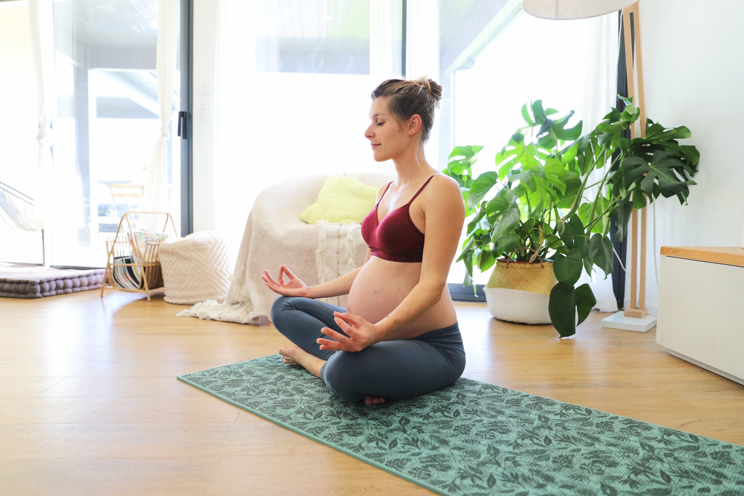 Femmes enceinte en tenue de sport qui médite sur un tapis de yoga et dans une ambiance zen et lumineuse avec des plantes